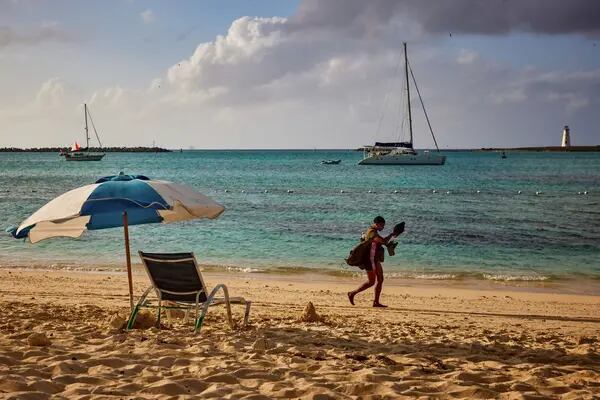 Pessoa passeando em uma praia. É possível ver barcos ao longe e, em primeiro plano, uma cadeira de praia e um guarda-sol