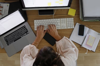Foto tirada de cima mostrando uma mulher digitando em um computador, ela também tem ao seu lado um laptop e um caderno com anotações
